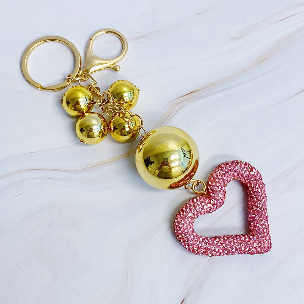 A gold-colored key chain featuring a series of round baubles with a pink heart-shaped charms with rhinestone embellishments.