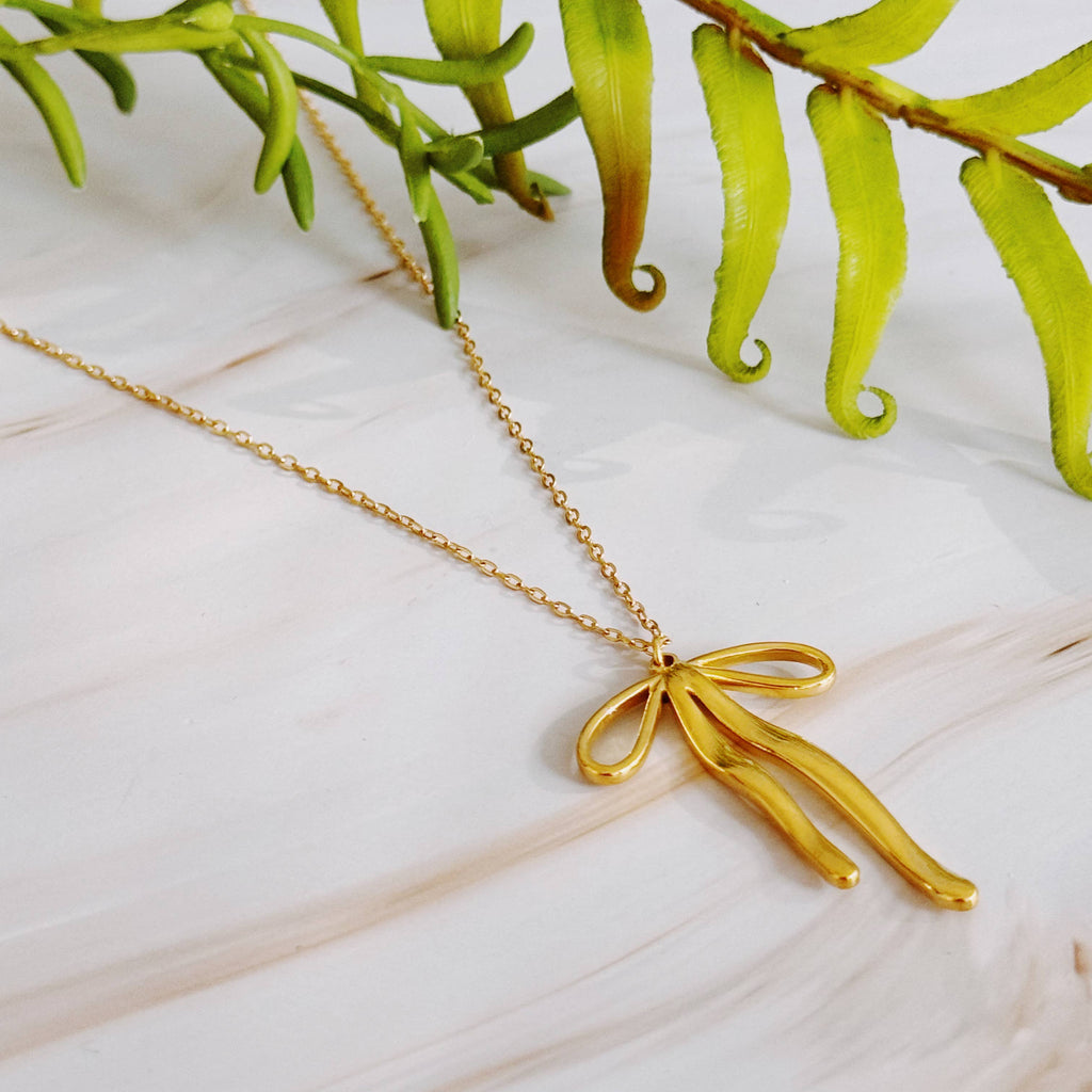 Gold necklace with a bow-shaped pendant on a light wooden surface with green leaves in the background.